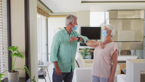 Senior-caucasian-couple-wearing-face-masks-greeting-each-other-by-touching-elbows-at-home