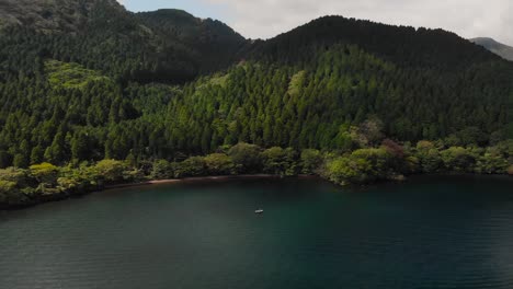 Slow-moving-backwards-aerial-drone-over-beautiful-lake-with-forest-and-single-fishing-boat-and-birds-flying