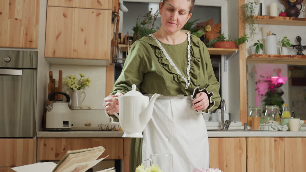 Housewife gently places white ceramic jug on table, smiles softly, sits down and rests chin on hand while admiring cupcake, surrounded by cozy kitchen elements and open book