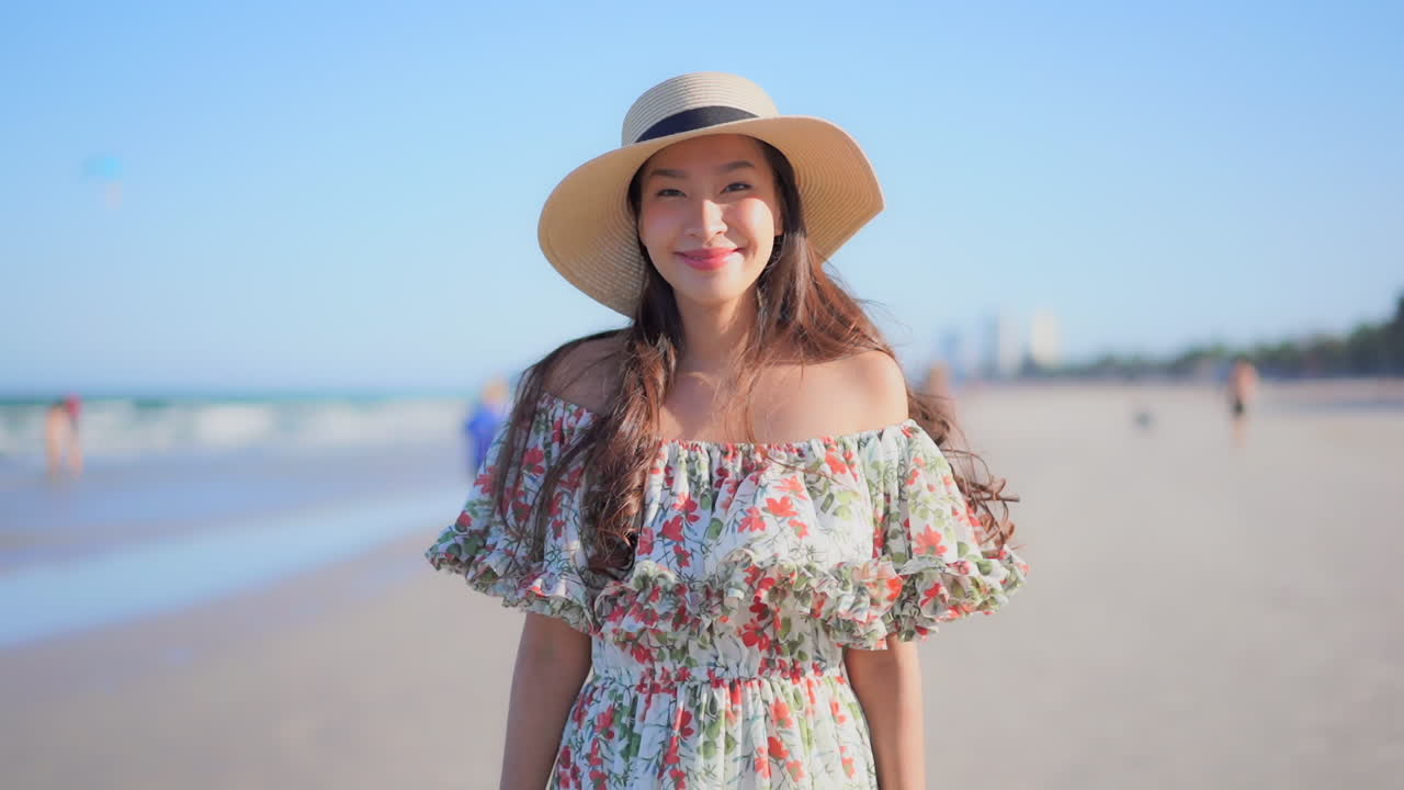 A medium close-up pretty young woman in a sundress and sun hat narrow depth of field beach Southeast Asia