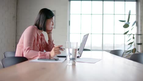 focused asian businesswoman using laptop and taking notes, at table in meeting room, in slow motion