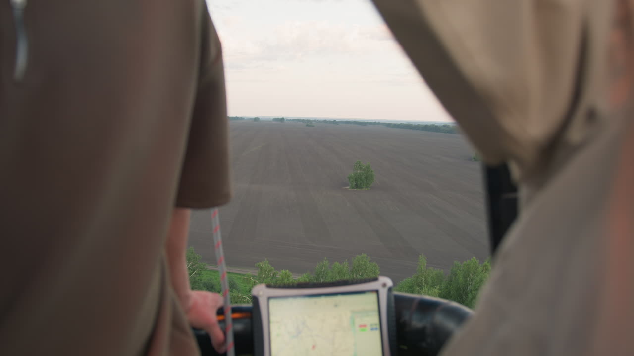 Overhead view from hot air balloon of vast tilled farmland with lone tree cluster on horizon, passengers holding basket edge and onboard tablet visible, capturing serene rural flight at dusk