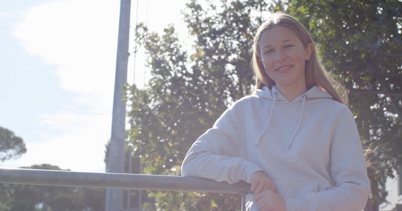 Adolescente con sudadera clara apoyada en una barandilla de hierro, el sol iluminando su cabello, haciendo que se gire y sonría.