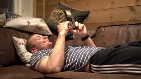 caucasian man laying on sofa and scrolling his phone while smiling and laughing - slow slider shot with focus pull and face in focus in end of shot