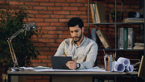 arabian businessman sitting at the table in the office, watching something on the tablet device screen and writing notes