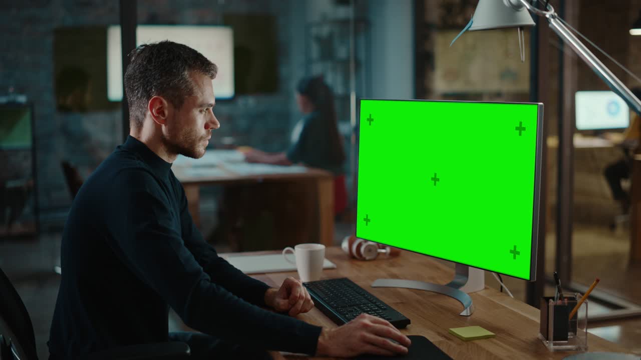 Young Handsome Specialist Working on Desktop Computer with Green Screen Mock Up Display in a Busy Creative Office with Colleagues. Male Manager with Trimmed Beard is Wearing a Casual Black Jumper.
