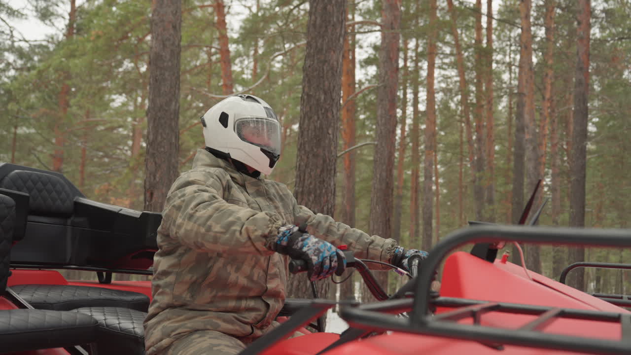 Pilota in mimetica su ATV nella foresta che percorre un sentiero tra i pini, controllando lo specchietto e poi afferrando il manubrio, accendendo il motore e guidando un quad rosso attraverso un terreno fangoso, casco integrale bianco e guanti visibili.
