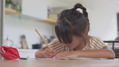 young asian girl home schooling working at table in kitchen writing in book during lockdown