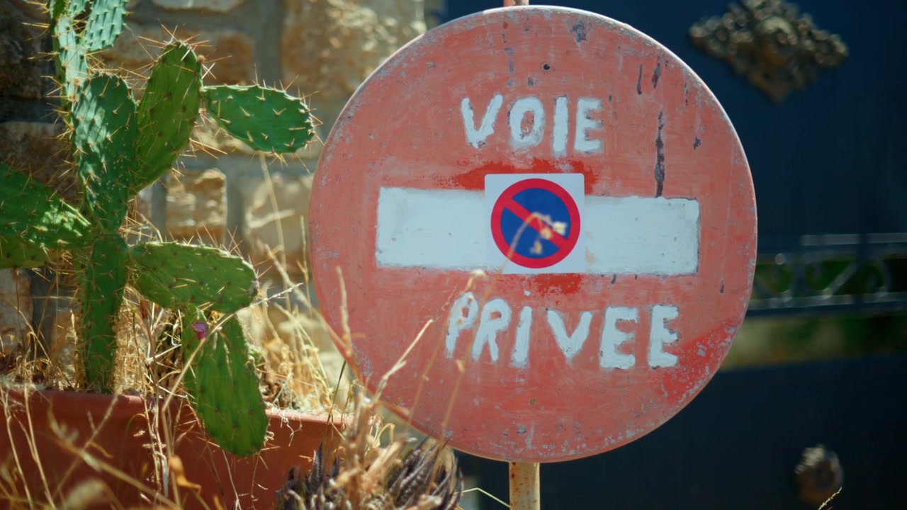 Gammelt, vejrbidt "Voie Privee" - privat vej - skilt med et parkeringsforbudssymbol, ved siden af en pottekaktus i solrige omgivelser.