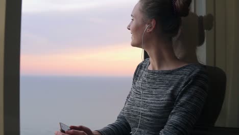 Niña-Sonriente-Parada-Junto-A-La-Ventana-Durante-Una-Puesta-De-Sol-Y-Escuchando-Música-Con-Auriculares-Usando-El-Teléfono-Móvil