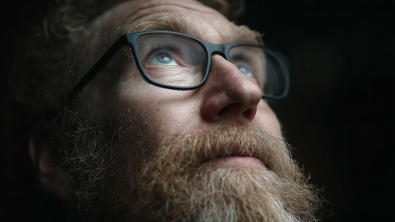 Close-Up Portrait of a Man with Glasses and Beard Contemplating in Low Light, Capturing Emotion and Depth in His Gaze
