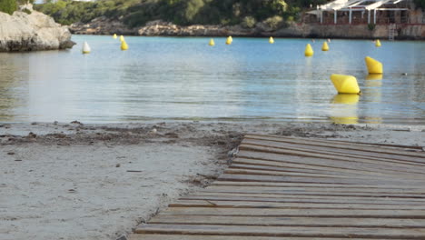 Cala-Santandria-in-Menorca:-sand,-wooden-path,-navigation-lane-of-yellow-buoys-indicating-the-way-for-boats,-Balearic-Islands,-Spain