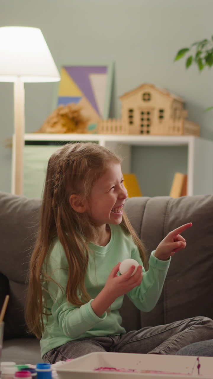 Little girl laughs and points by finger to elder sister holding Easter egg during coloring at home slow motion. Ebru art technique usage in decor