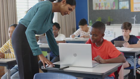 teacher helping student with laptop in classroom, other students studying