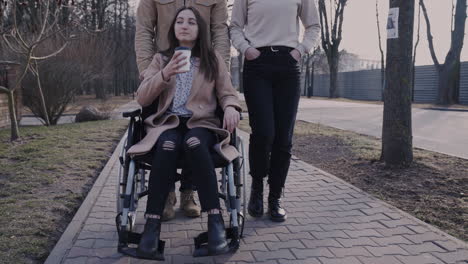 Front-View-Of-Woman-And-Man-Taking-A-Walk-With-Her-Disable-Friend-In-Wheelchair-Around-The-City-1