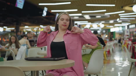 lady in pink closes her laptop, looking bored and tired in a busy restaurant as she takes a deep breath and stretches, other people are seated in the restaurant, with a slight blur in the background