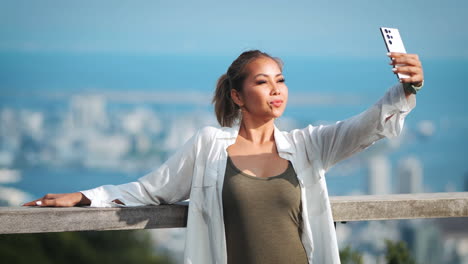 Young-female-model-takes-selfie-with-a-white-smartphone,-framed-by-a-cityscape-and-clear-blue-sky