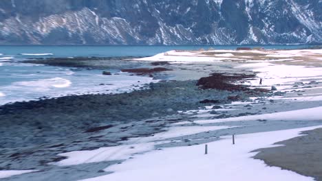 Zoom-back-from-hikers-walking-to-reveal-waves-roll-into-a-beautiful-snow-covered-shoreline-amidst-fjords-north-of-the-Arctic-Circle-in-Lofoten-Islands-Norway