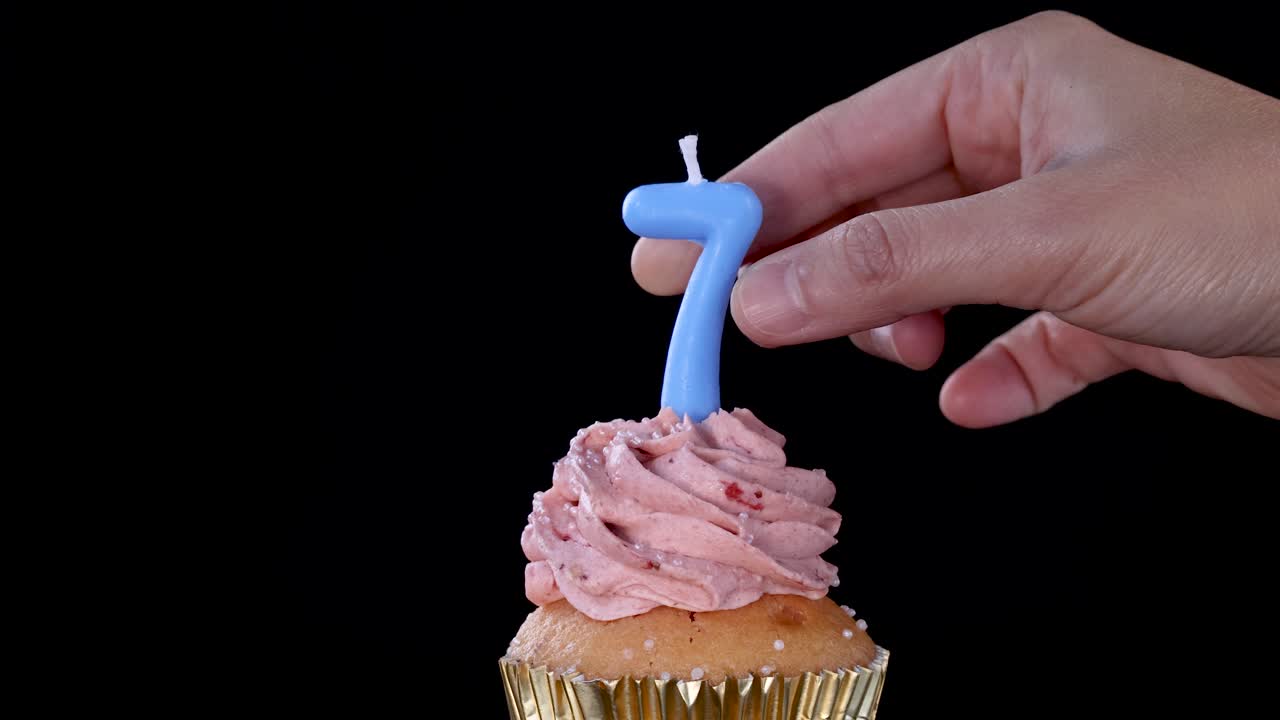 A hand inserts a blue number seven birthday candle into a cupcake topped with pink strawberry frosting against a black background, using steady close-up framing
