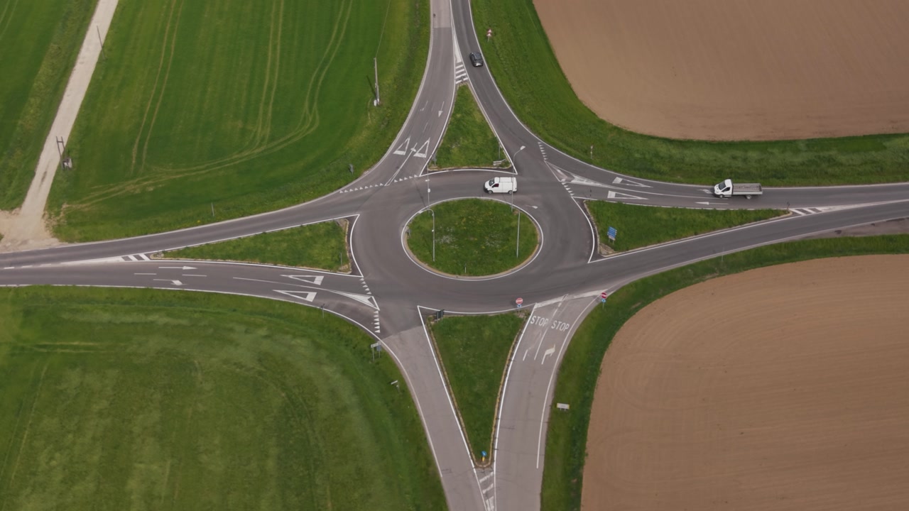 Drone aerial view of a roundabout surrounded by rural fields and countryside. Vehicles move through the intersection, blending infrastructure with natural landscapes.