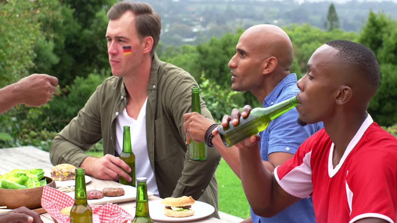 Amici di diverse etnie a un tavolo da picnic che tengono in mano bottiglie di birra, reagendo a una mano che indica fuori campo.