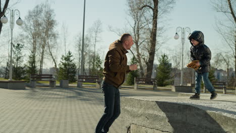 a father and son playing together outdoor as the son moves forward and removed his beanie playfully, with trees in the background