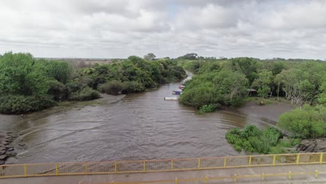 the drone follows the winding river through dense greenery in punta lara, buenos aires, capturing the natural landscape with boats along the riverbank.