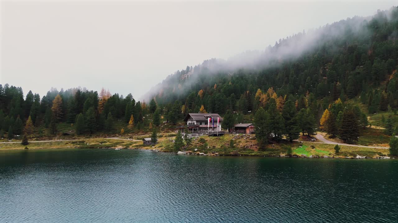 Luftoptagelse af en bjerghytte nær Oberseesøen, omgivet af en tåget skov ved Staller Sattel. Efterårslandskabet med farverige træer og stille vand ved grænsen mellem Østrig og Italien.