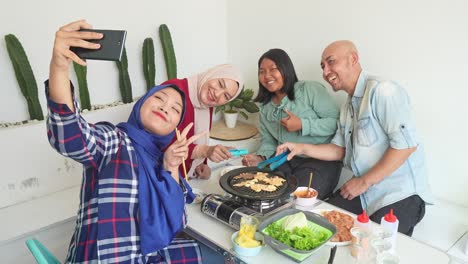 cheerful asian muslim family, mother, father and daghters, taking selfie using smartphone while eating korean bbq together