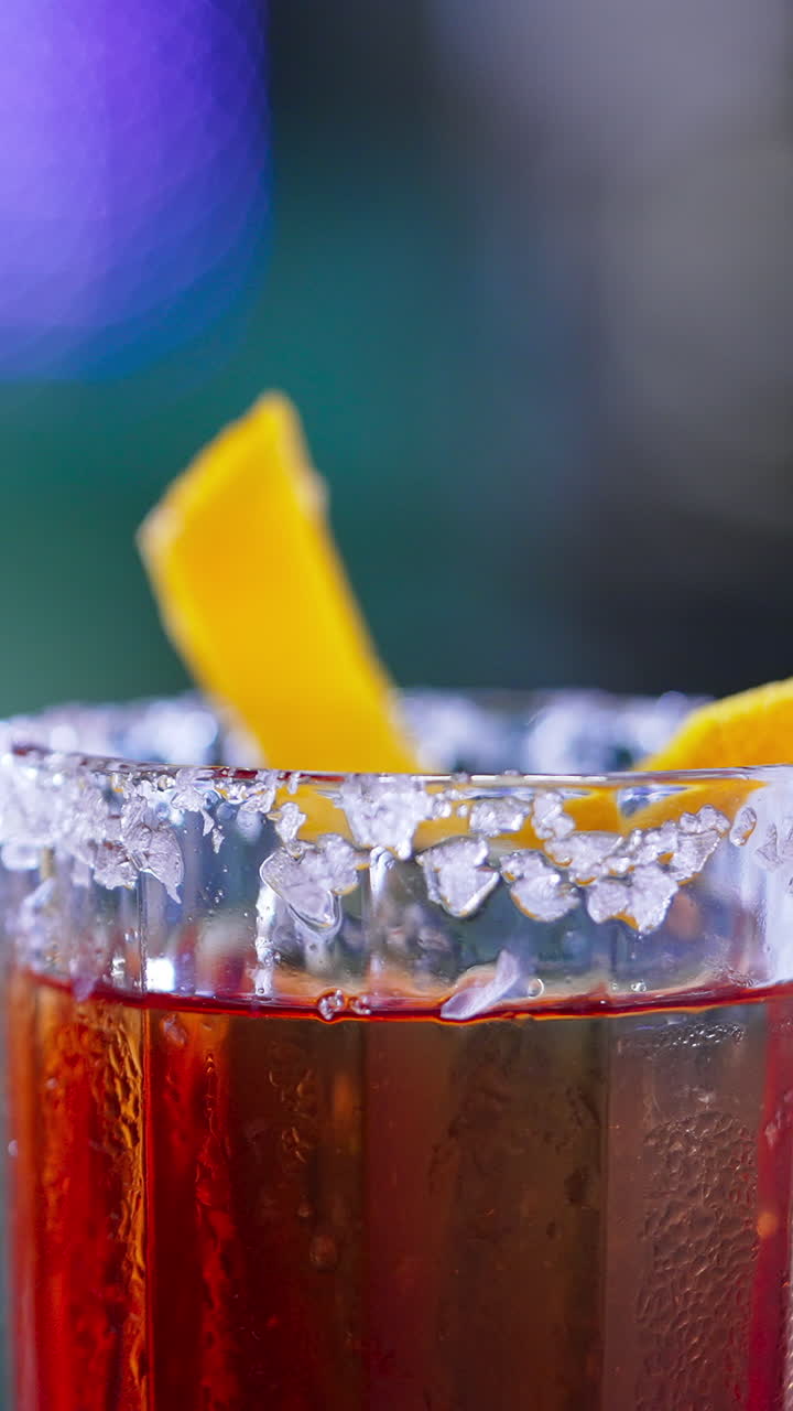Rotating glass with top decorated with salt and orange peel. Red alcohol cocktail in the bar. Close up. Blurred backdrop. Vertical video