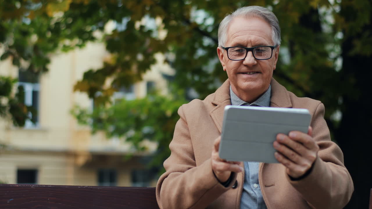 anciano con gafas y abrigo sentado en el banco del parque y usando una tableta en otoño