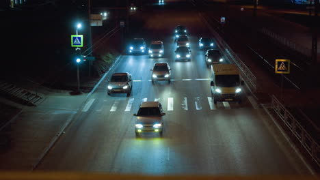 close view of nighttime traffic at a pedestrian crossing with multiple cars and their headlights illuminating the road, streetlights and pedestrian signs are visible