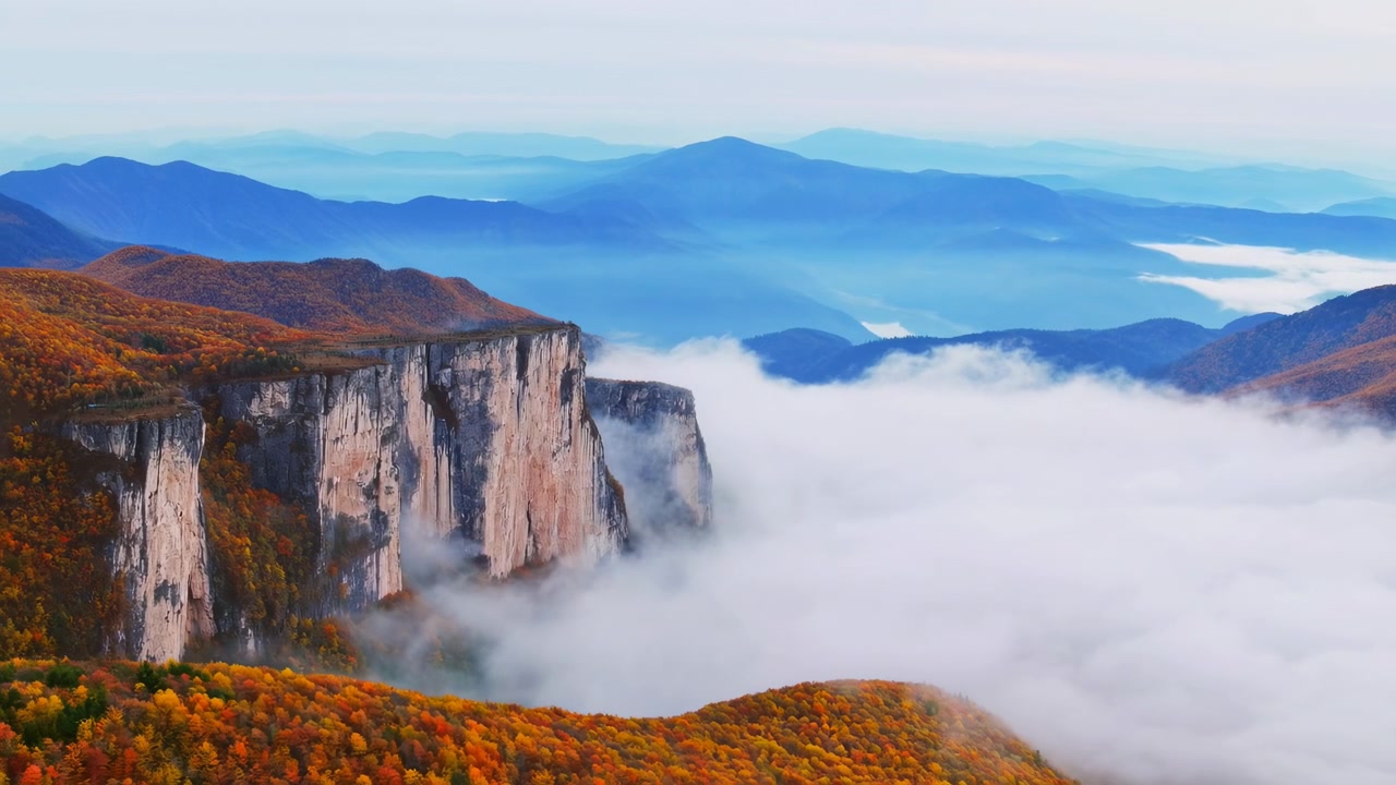 Una nebbia bianca si sposta lentamente attorno ad alte scarpate di pietra e pendii boscosi dai caldi colori autunnali, rivelando silhouette montane a strati sotto una pallida luce naturale del giorno.