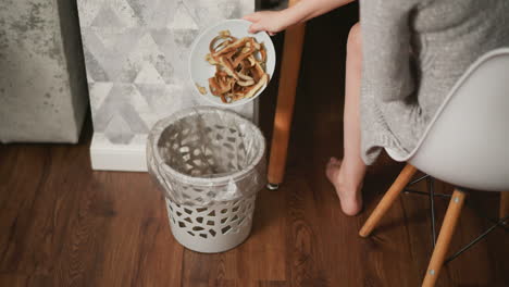woman seated on white chair disposing potato peels into plastic-lined dustbin, bare feet resting on polished wooden floor, cozy home setting with patterned wall and nearby cushioned sofa visible
