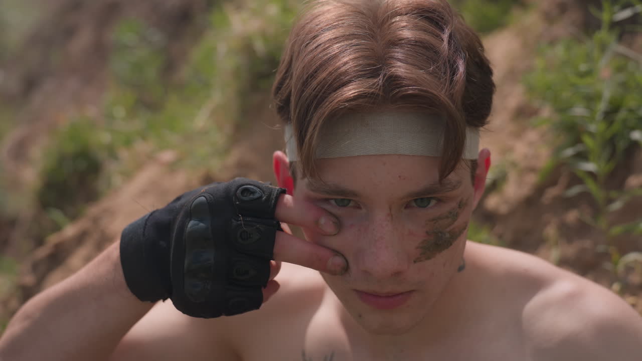 Shirtless adventurer with stern expression sits near grassy riverbank, face smeared with sand, head wrapped in beige band, chest tattoo visible, sun casting soft shadows on skin, moment of focus