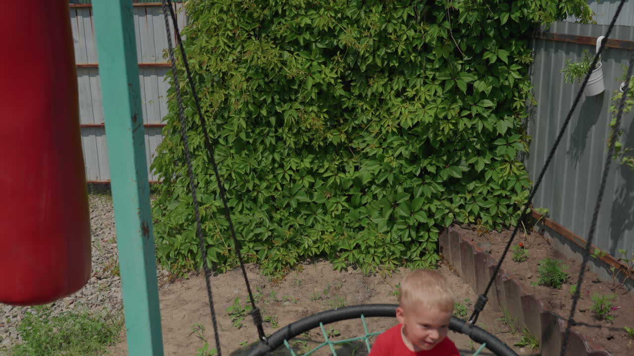 Caucasian Boy Helping Friend Into Nest Swing In Backyard, Steadying Rope And Guiding Feet, Sunny Day With Green Hedge And Punching Bag Nearby, Small Act Of Cooperation And Playful Teamwork