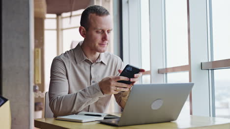 handsome smiling man sitting at the desk in front of laptop. male employee takes his phones and checks the information there.