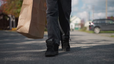 close-up of legs walking in residential area, person carrying paper bag, with autumn trees, houses, and a parked car in the background, capturing a suburban autumn day