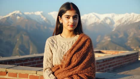 young woman wearing a cozy beige cable knit sweater and a stylish brown shawl, posing against a stunning snowy mountain range on a bright, sunny winter day