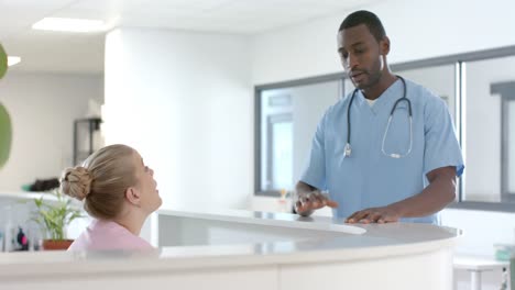 African-american-male-doctor-talking-to-medical-receptionist-at-front-desk-at-hospital,-slow-motion
