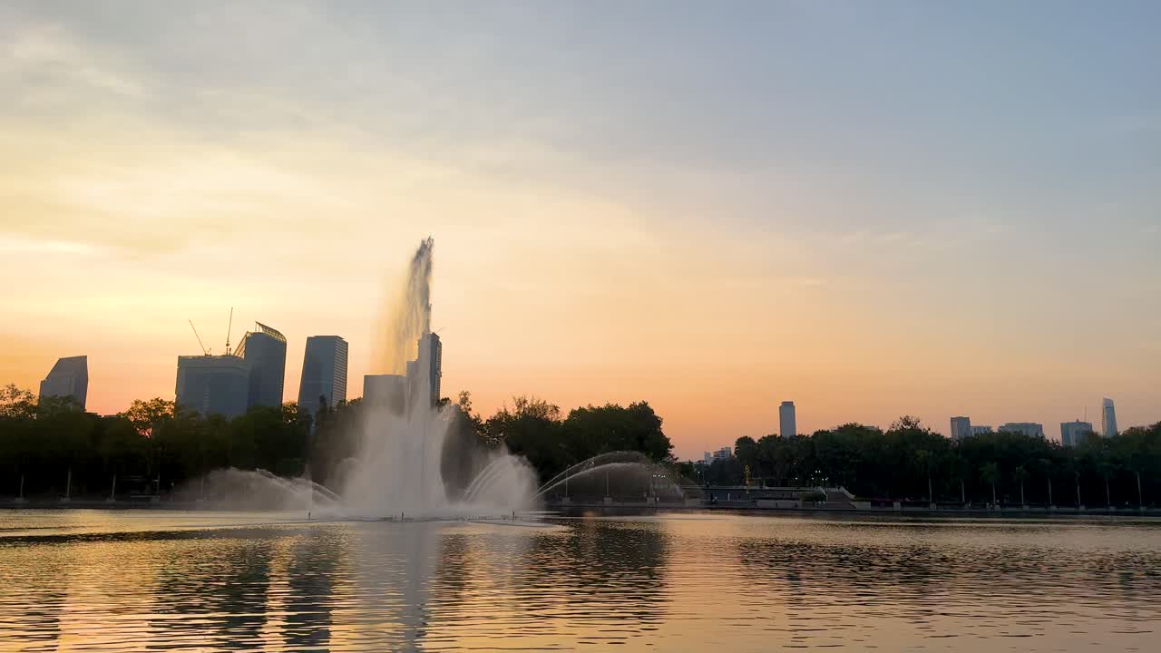 A serene sunset scene at Benjakitti Park with a fountain, city skyline, and vibrant foliage