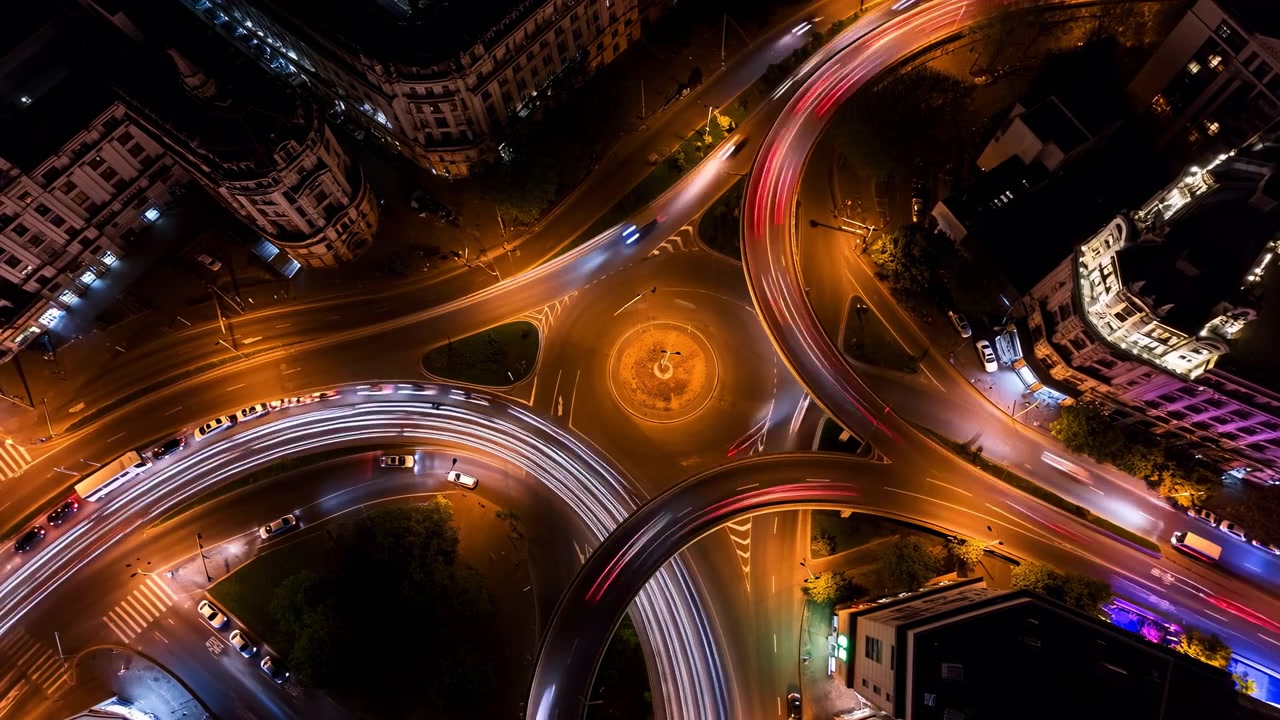 Il traffico notturno dall'alto si muove lungo strade curve e ampie attorno a un'isola centrale illuminata, formando scie luminose bianche e rosse tra gli edifici circostanti e la fitta copertura arborea