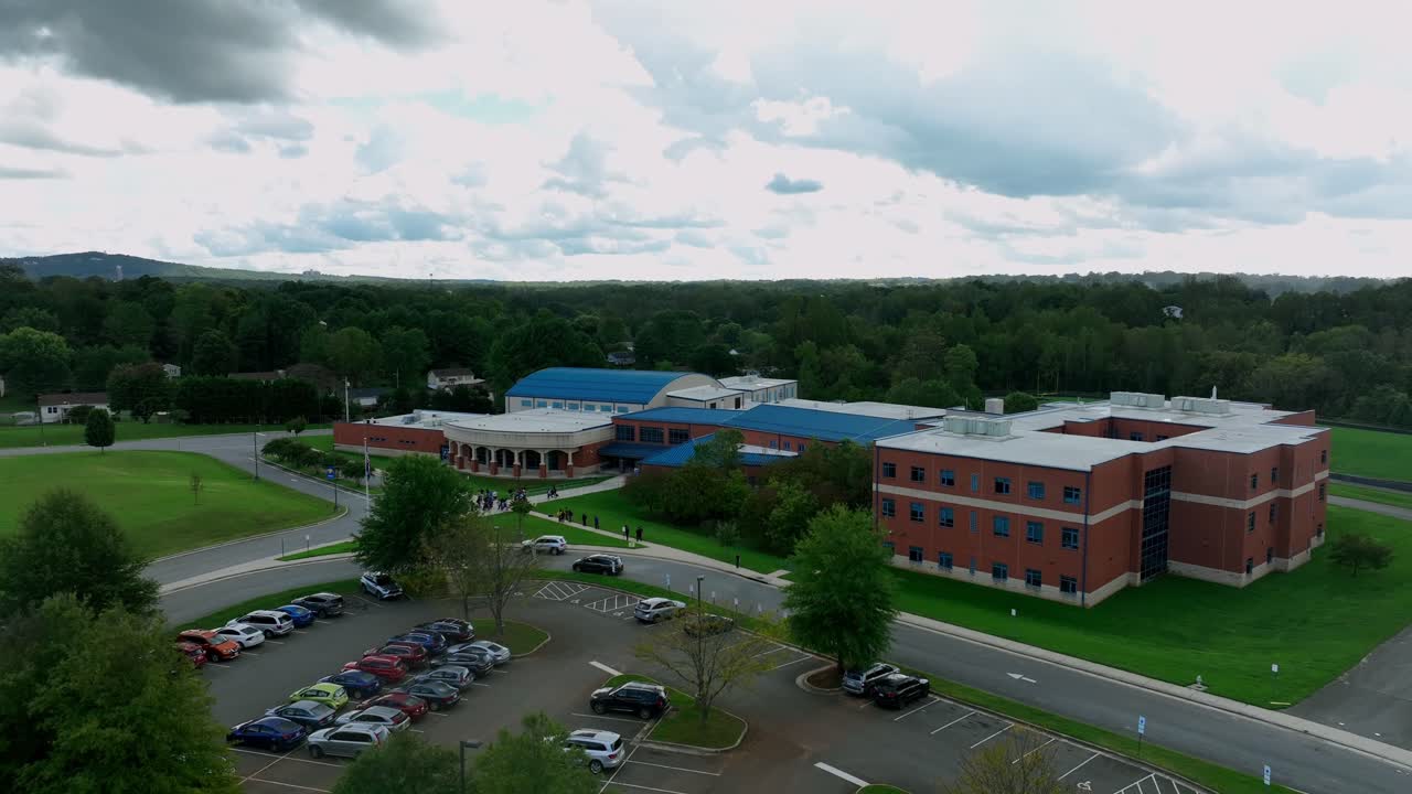 Aerial descend shot of parking cars in front of American elementary school with red bricks. Waiting pupils on sidewalk. Cloudy day with green trees in autumn season. Wide shot.