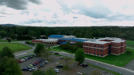 aerial descend shot of parking cars in front of american elementary school with red bricks. waiting pupils on sidewalk. cloudy day with green trees in autumn season. wide shot.