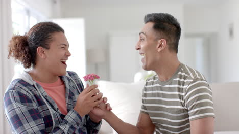 happy diverse gay male couple sitting on couch and giving flower in living room, slow motion