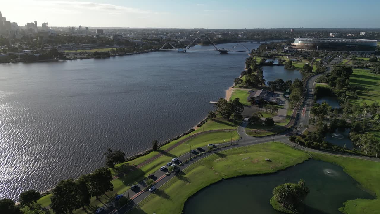 vuelo aéreo de drones sobre el lago, carretera costera del río swan en la ciudad de perth durante un día soleado - puente matagarup y estadio optus en el fondo