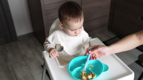 baby boy in white shirt sits in front of a blue plate. kid holding a spoon. mom feeding a son. top view.