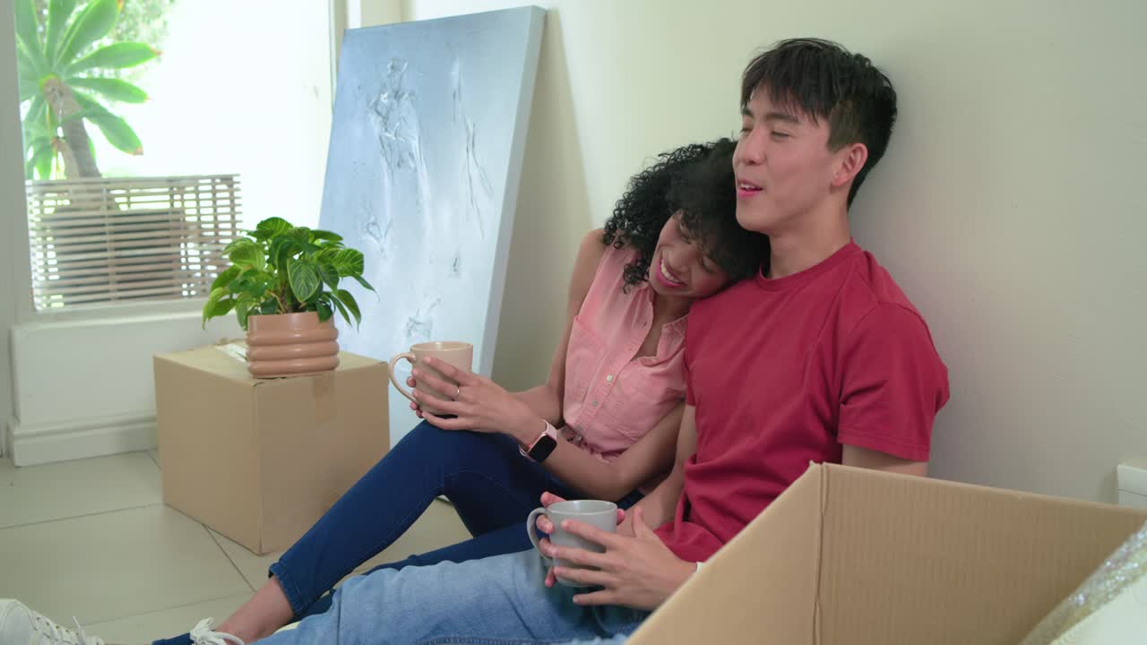 荷ほどきをしながら引っ越し箱に囲まれて座っている多様なカップル。マグカップを手に談笑し、肩にもたれかかっている。