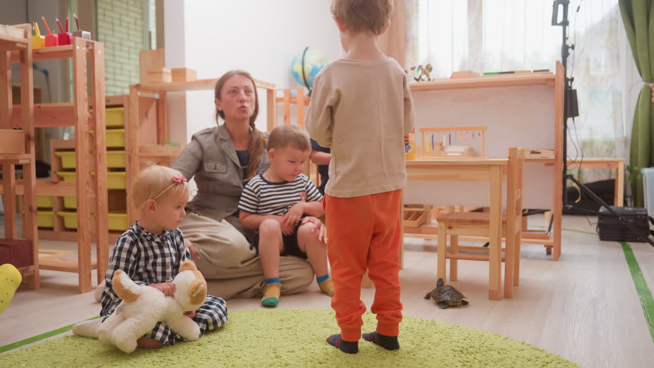 Tortoise walking towards chair in classroom while mother carries kid interacting with another boy standing aside as little girl sits holding stuffed toy observing scene with curiosity