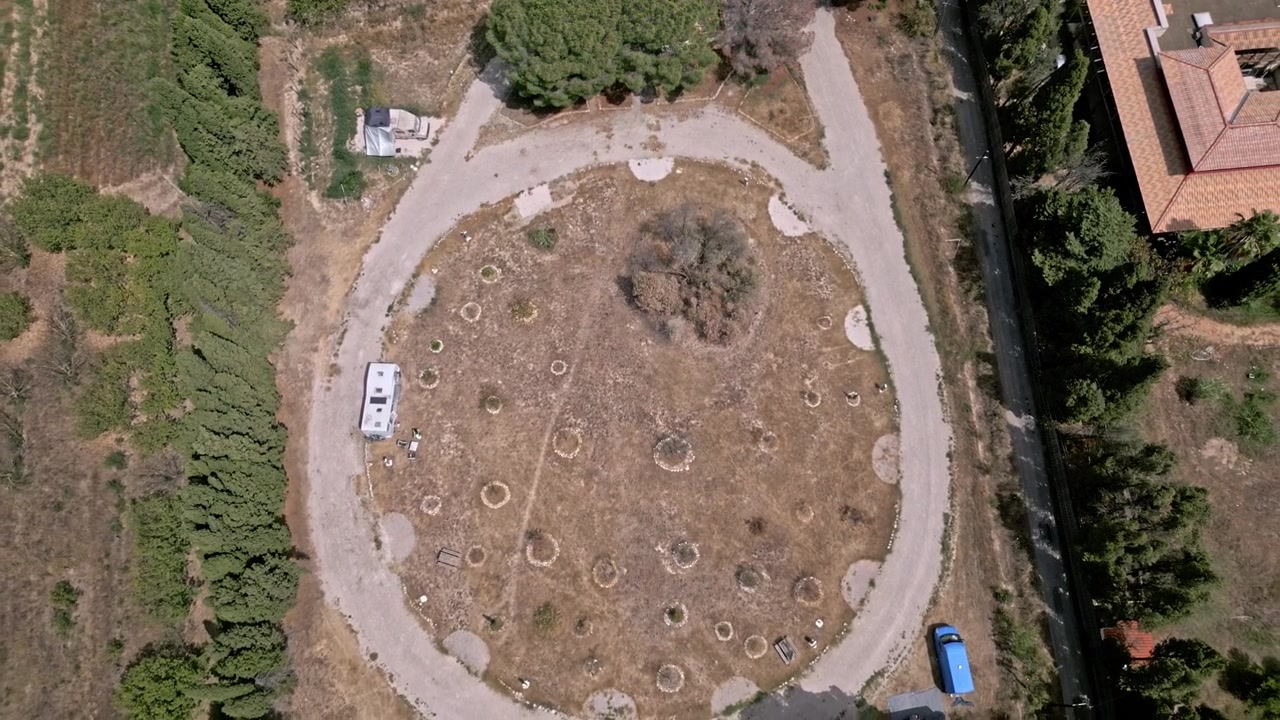 Vista aerea di un camper bianco parcheggiato su un'area circolare sterrata all'interno di un campeggio. Una casa con tetto rosso è visibile nelle vicinanze.